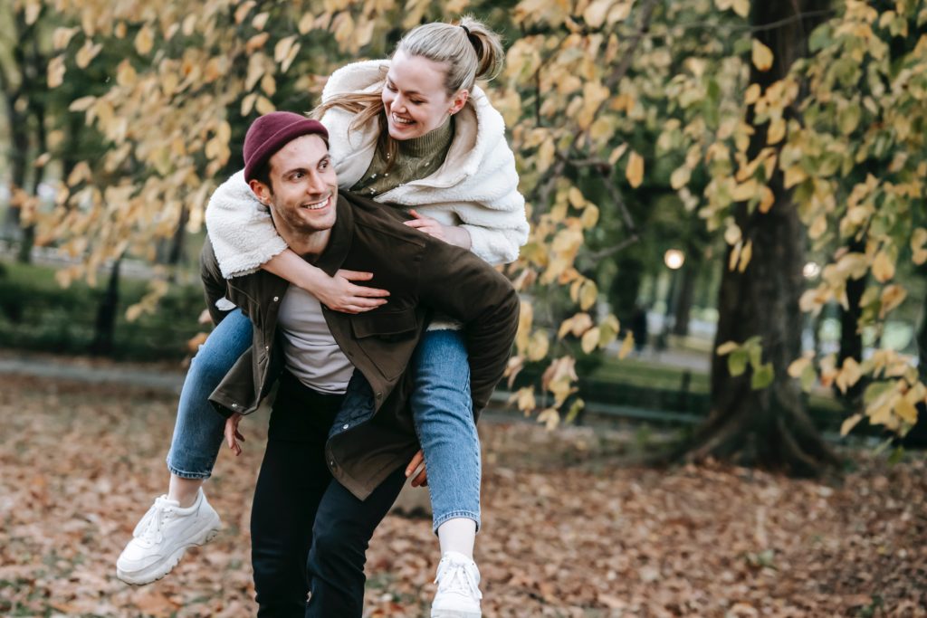 A man is giving a woman a piggyback ride against an autumnal background.
