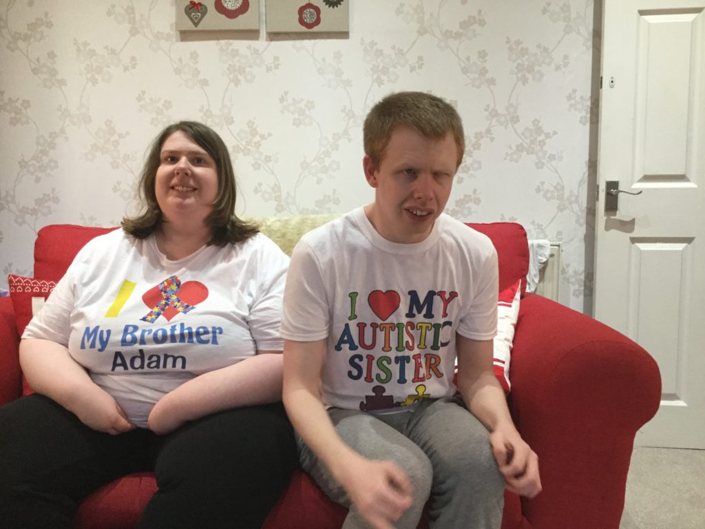 April Slocombe and her brother, Adam, are sitting on a couch. They are both wearing autistic sibling-themed T-shirts.