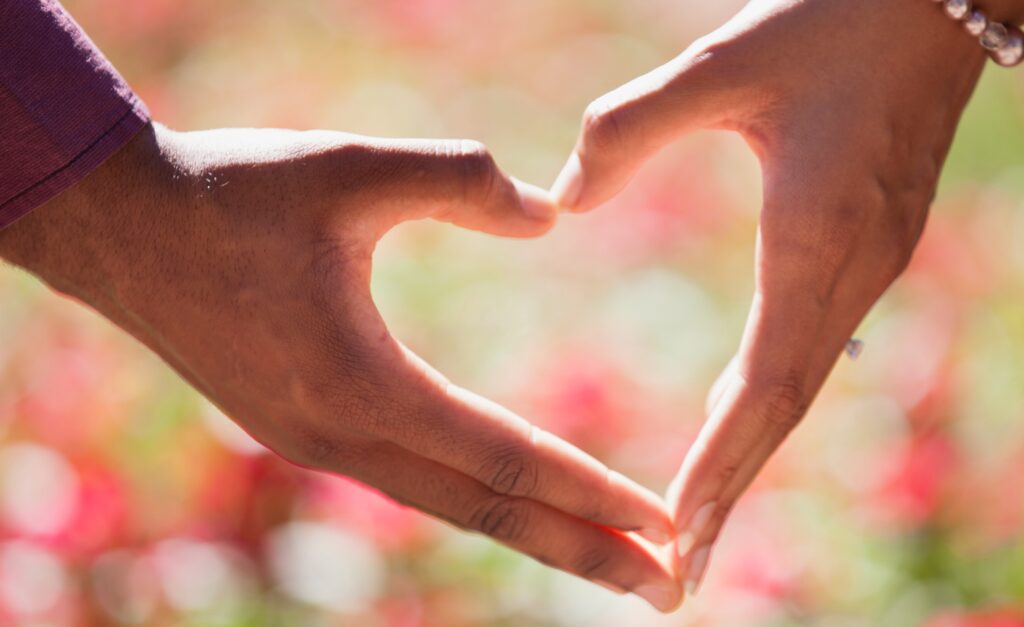 Two hands form a heart shape against a blurred floral background.