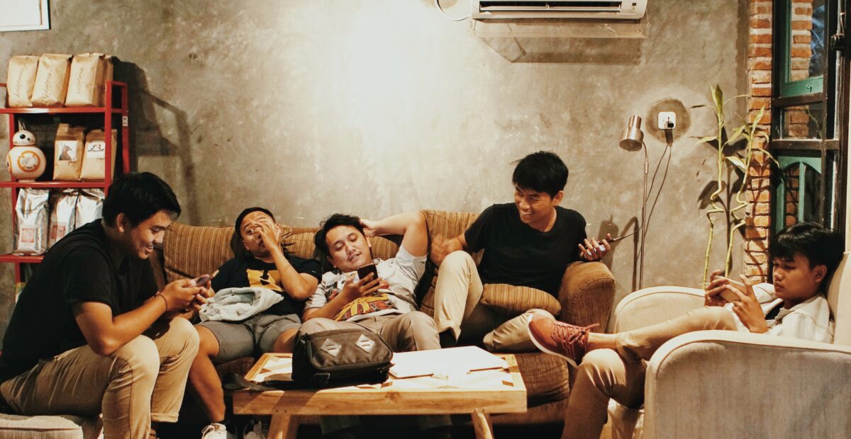 Five people are sitting on armchairs and a sofa around a coffee table. Three people are using their phones, one person is looking at another and one person looks anxious.