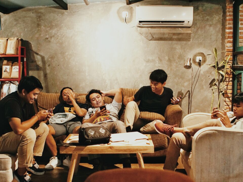 Five people are sitting on armchairs and a sofa around a coffee table. Three people are using their phones, one person is looking at another and one person looks anxious.