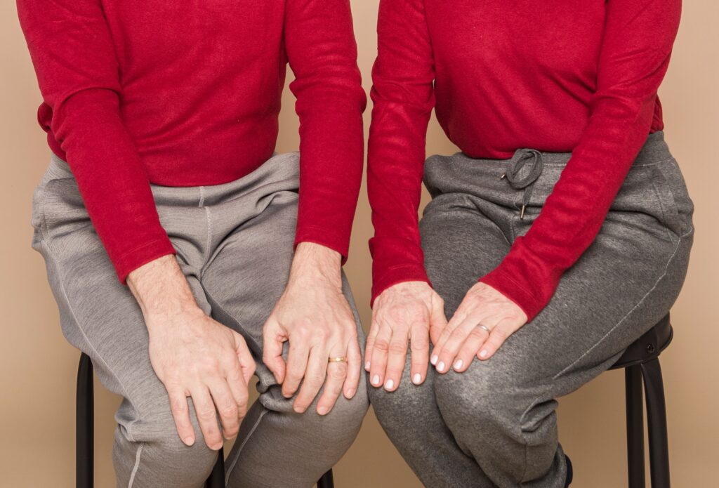 A couple dressed in red jumpers and grey trousers are sitting on stools.