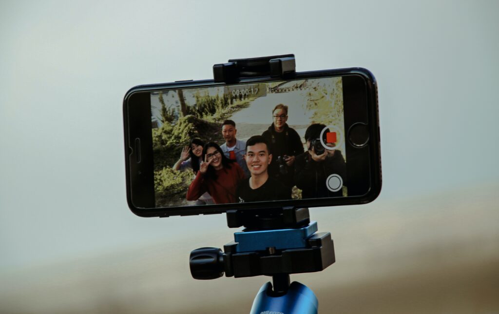 A smartphone is mounted on a tripod. The smartphone is recording a video of a group of people smiling and waving.