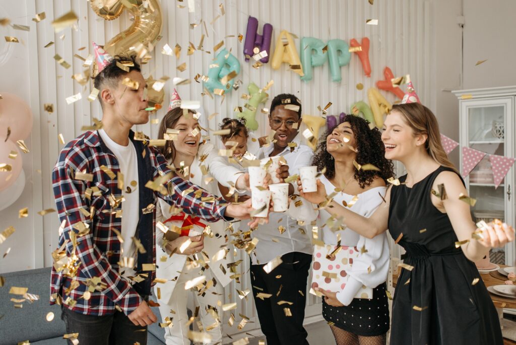 Six people are saying, "cheers," with their plastic cups at a birthday party. There is confetti in the foreground.