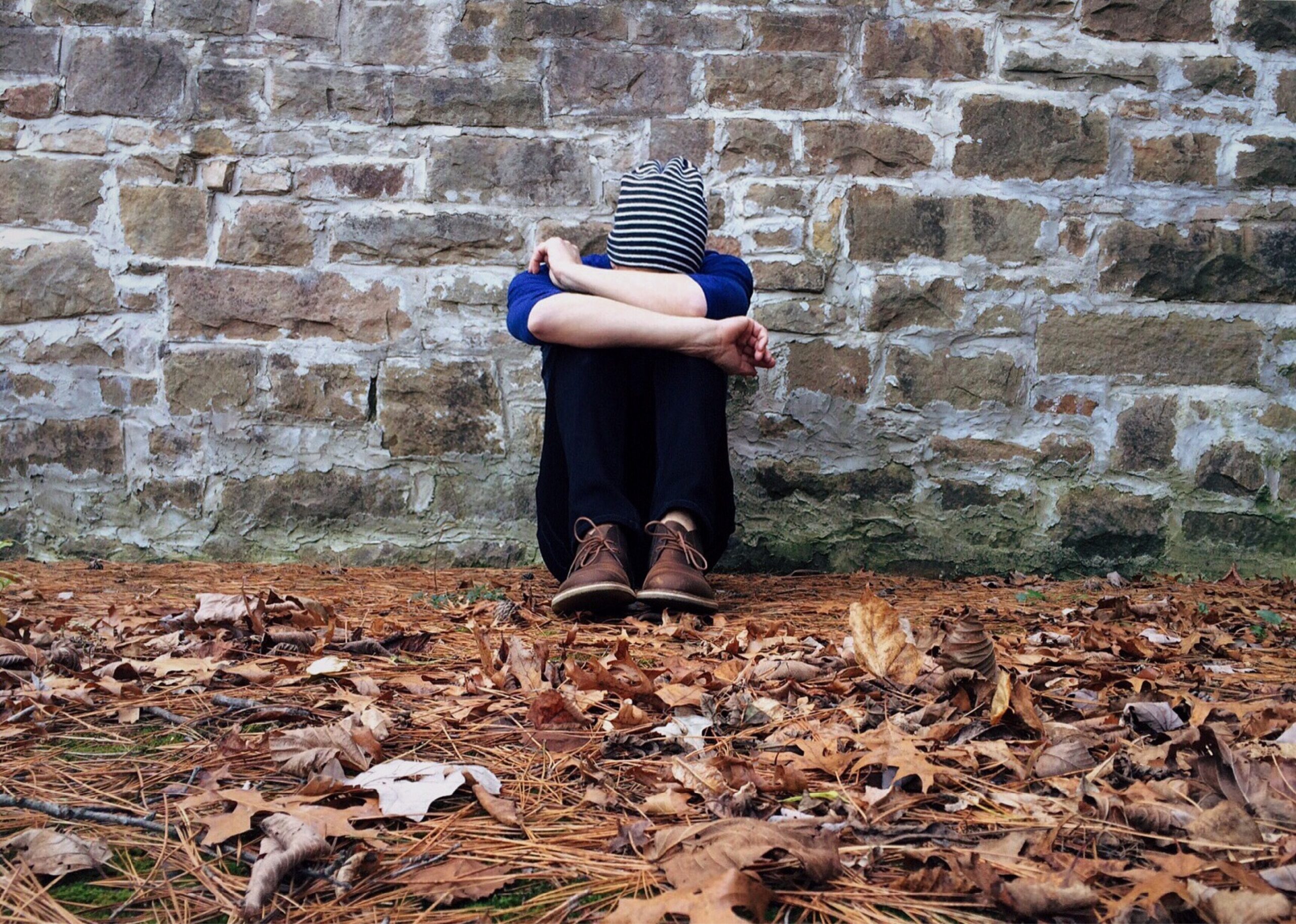 A person is sitting against a brick wall. Fallen leaves lie on the ground. The person has their head buried in their arms and their arms crossed.