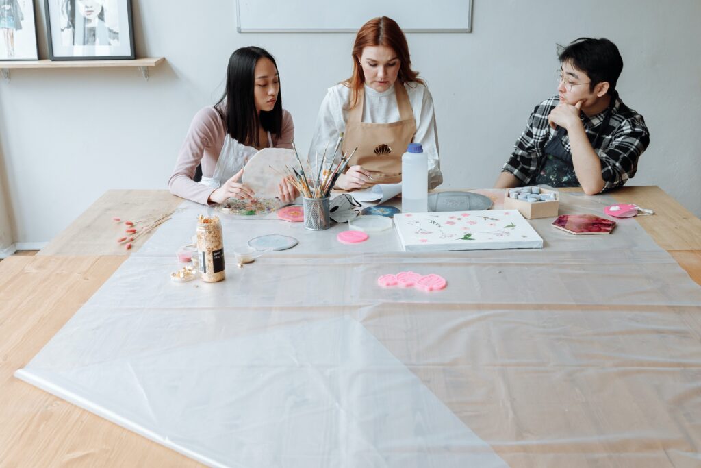 Three people are sitting at a table. One person is painting and the other two people watch her. A canvas with a floral print lies on the table.