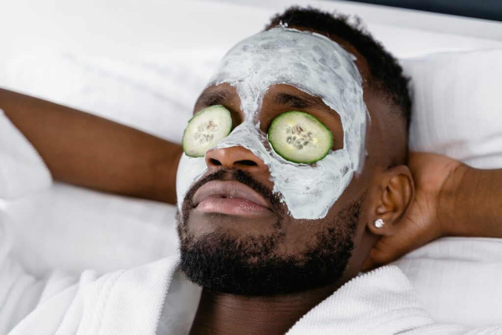 A close-up of a man with a clay mask on his face and cucumber slices on his eyes.
