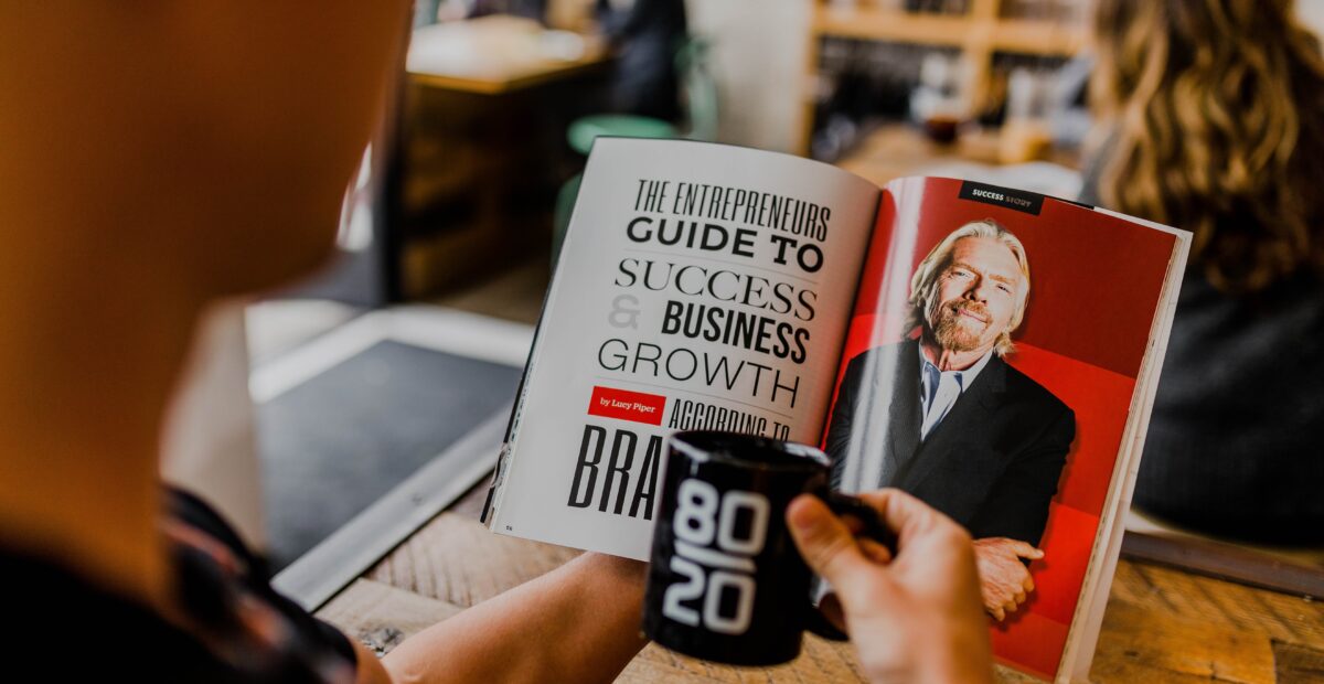 A person is holding an open book in their left hand. The book has text on the right page and a photograph of Richard Branson on the other page. The person is holding a mug in their other hand.