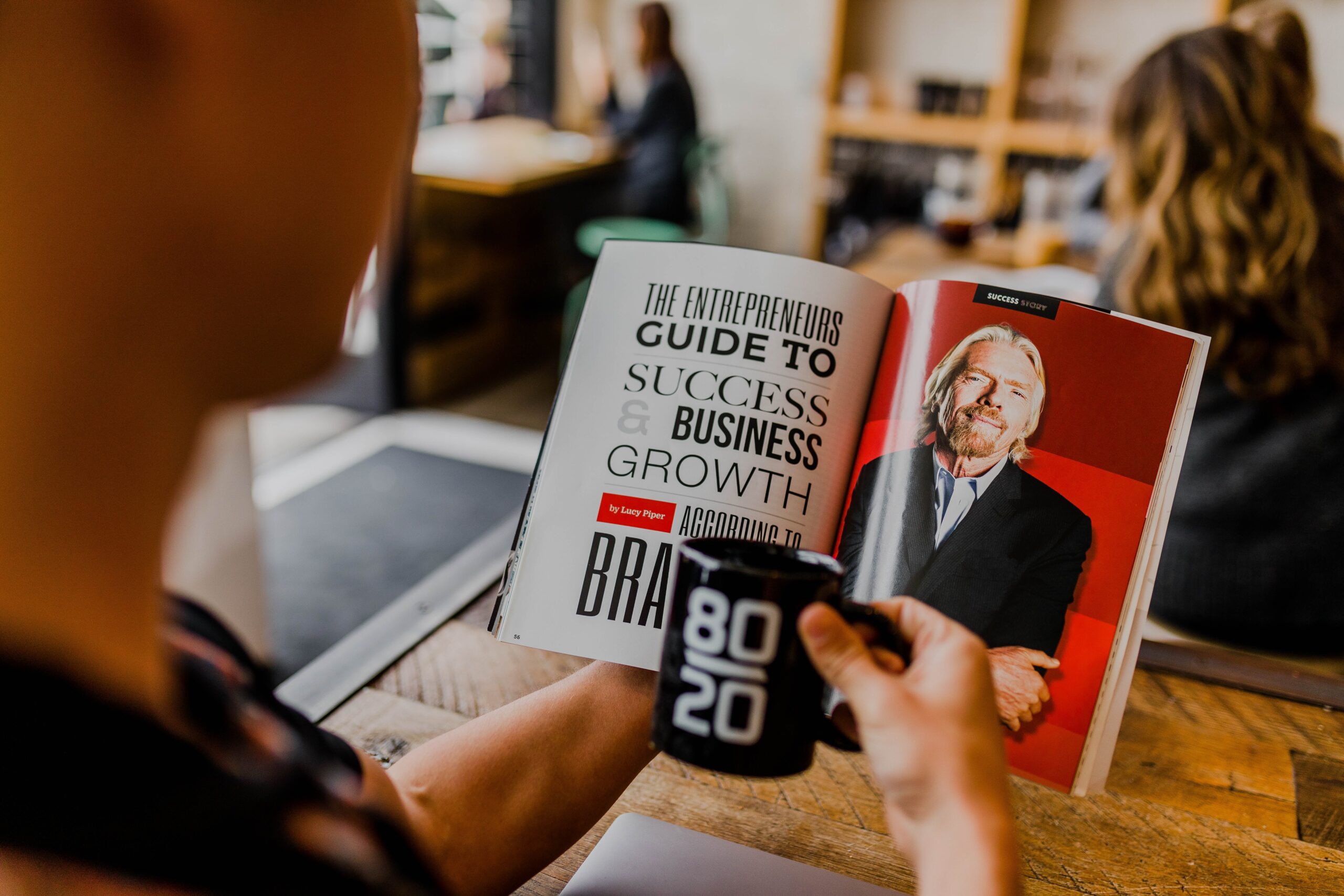 A person is holding an open book in their left hand. The book has text on the right page and a photograph of Richard Branson on the other page. The person is holding a mug in their other hand.