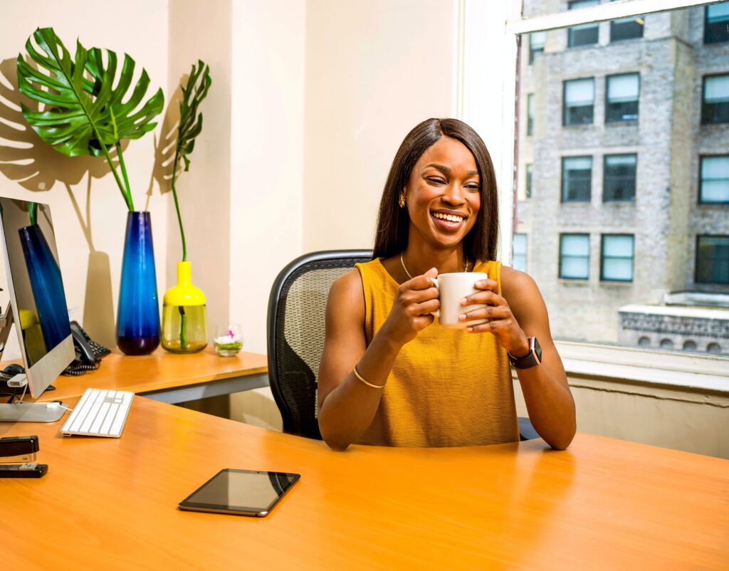 A woman smiles as she sits at her desk and holds a coffee mug.